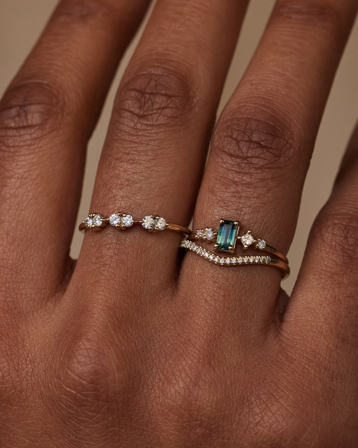 [lifestyle]Close-up of a hand wearing two diamond rings with a green gemstone on a neutral background