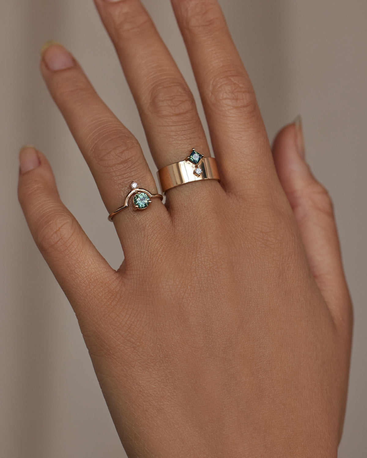 [lifestyle]Hand wearing two gold rings with green gemstones on a neutral background