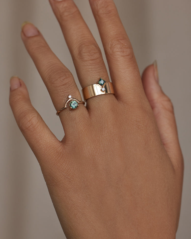 [lifestyle]Hand wearing two gold rings with green gemstones on a neutral background